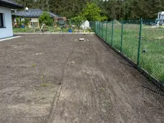 Przygotowanie terenu pod nowe nasadzenia. Widać świeżo zaoraną ziemię obok domu i ogrodzenia.