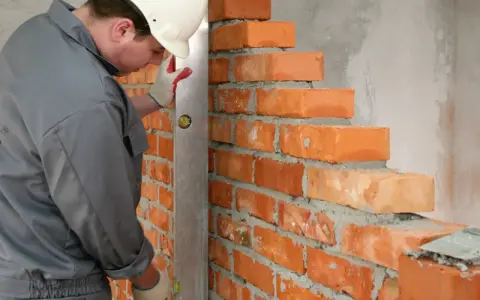 Murarz w kasku i rękawicach sprawdza pion muru. Cegły o standardowych wymiarach układane są na zaprawie.