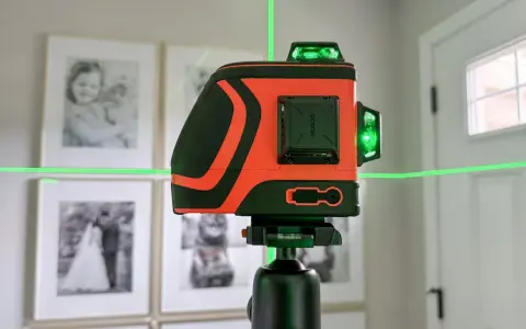 Pomarańczowa poziomica laserowa z zielonymi liniami na statywie, na tle oprawionych zdjęć.