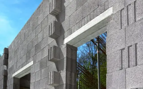 Ściana z bloczków betonowych, widoczny betonowy nadproże nad otworem okiennym. W tle zbrojenie i drzewa.