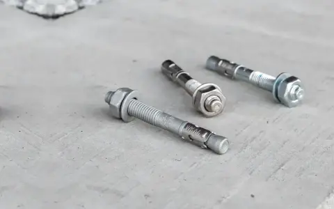 Trzy metalowe kotwy leżą na betonowej powierzchni. Każda kotwa ma gwintowany pręt i nakrętkę.