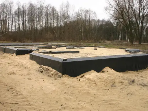 Fundament domu w piasku, otoczony czarną folią. W tle las. To początek budowy, gdzie dysperbent chroni fundament.