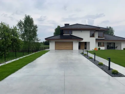 Nowoczesny dom z drewnianą bramą garażową, otoczony zielenią. Szeroki podjazd betonowy prowadzi do wejścia, a wzdłuż niego biegną lampy i rabaty z kamieniami.