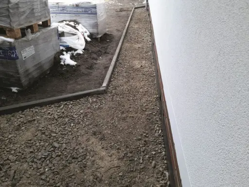 Budowa opaski wokół domu z kamienia i obrzeży. W tle palety z materiałami budowlanymi.