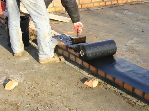 Pracownik nakłada masę asfaltowo kauczukową na mur ceglany, przygotowując podkład pod hydroizolację.