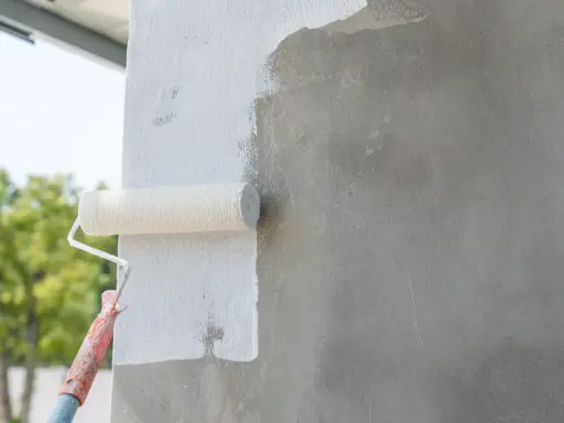 Malowanie betonu wałkiem. Biała farba pokrywa szarą ścianę, nadając jej świeży wygląd.