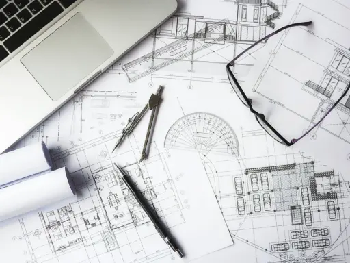 Na stole architekta leżą plany budynku, laptop, okulary i cyrkiel. To miejsce pracy nad projektem i jego kosztorysem.