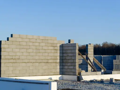 Budowa domu w toku. Wznoszona jest ściana nośna z bloczków betonowych, widoczne fundamenty i materiały budowlane.