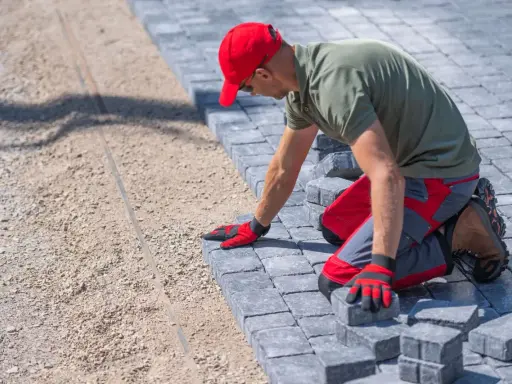 Pracownik w czerwonej czapce układa kostkę bauma na podsypce.
