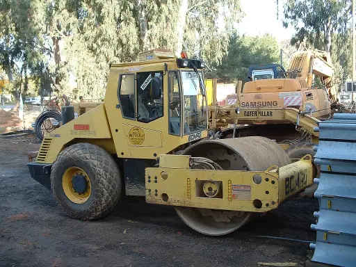 Żółty walec drogowy BOMAG BW 213D-3 na placu budowy, obok koparki Samsung Caterpillar.