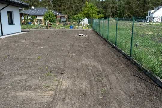 Przygotowanie terenu pod nowe nasadzenia. Widać świeżo zaoraną ziemię obok domu i ogrodzenia.